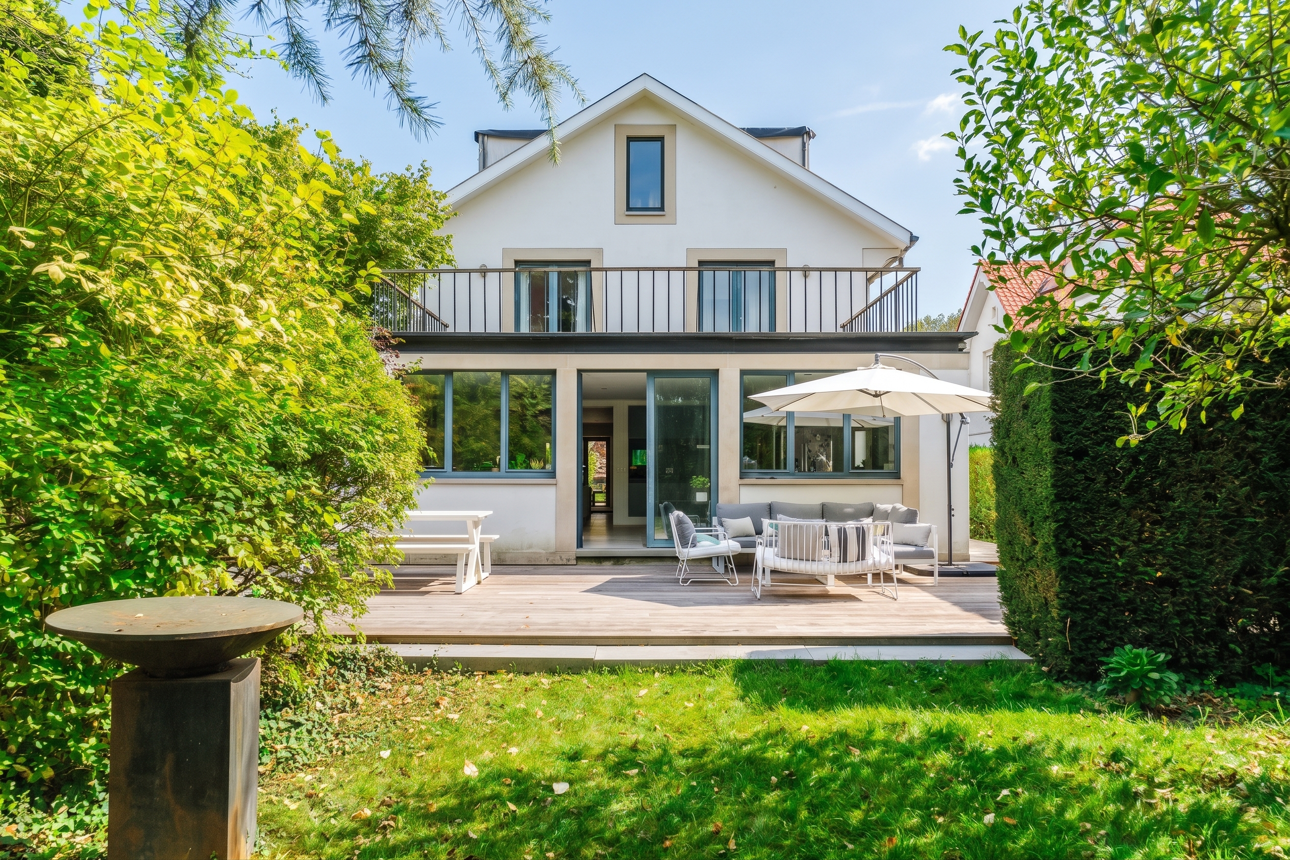 White house with a wooden deck, patio furniture, umbrella, and lush green garden on a sunny day. [AI-generated text] [Modifié par IA]