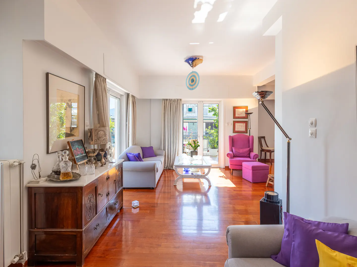 Bright living room with hardwood floors, a grey sofa, a vibrant pink armchair, and a wooden sideboard with decor.