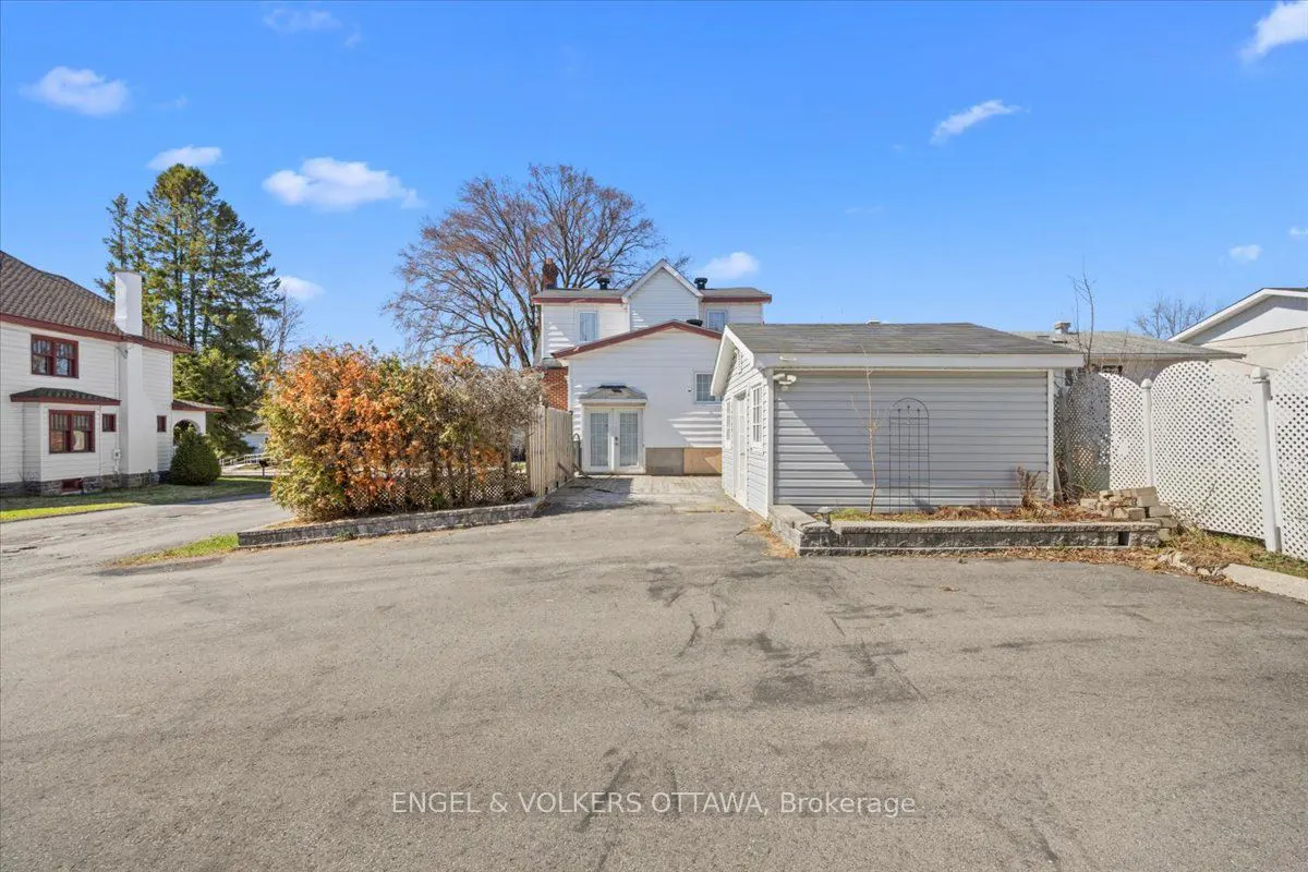 Backyard view of a white house with a gray garage, a hedge, and a paved driveway under a blue sky.
