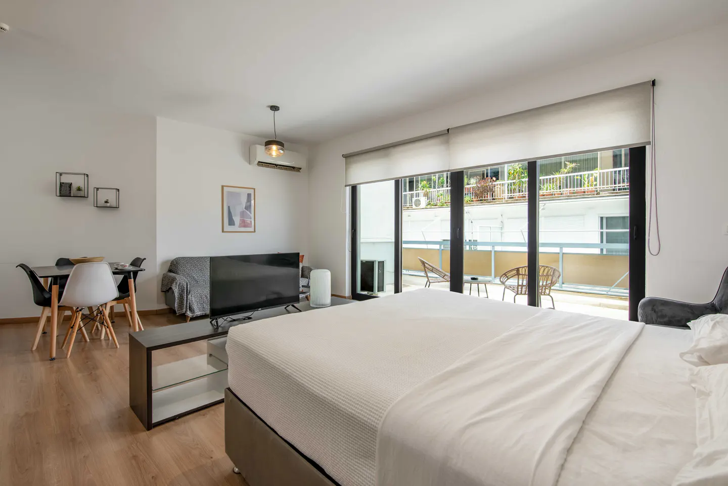 Bright studio apartment with a bed, TV, dining table, and balcony with chairs. The walls are white and the floor is wood.