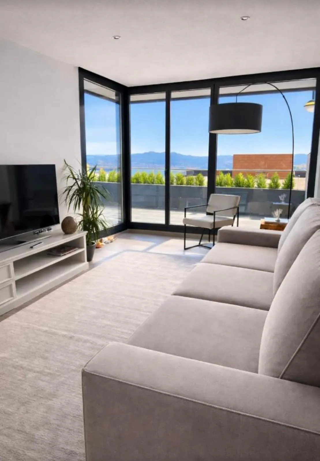 Modern living room with a beige sofa, TV on a white stand, potted plant, and floor-to-ceiling windows with a mountain view.