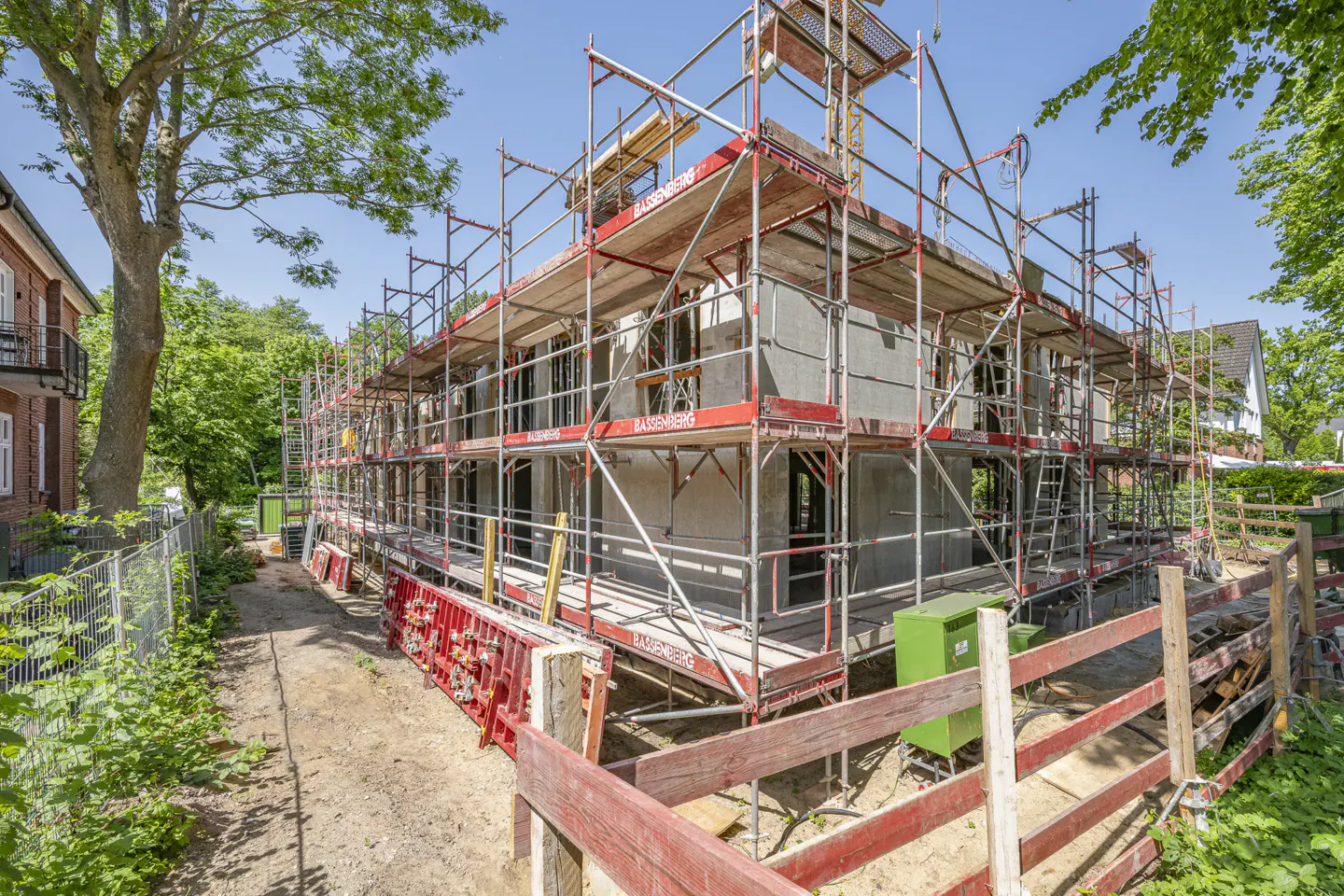 Exterior view of a building under construction, surrounded by red scaffolding and a wooden fence. Trees and blue sky in the background.