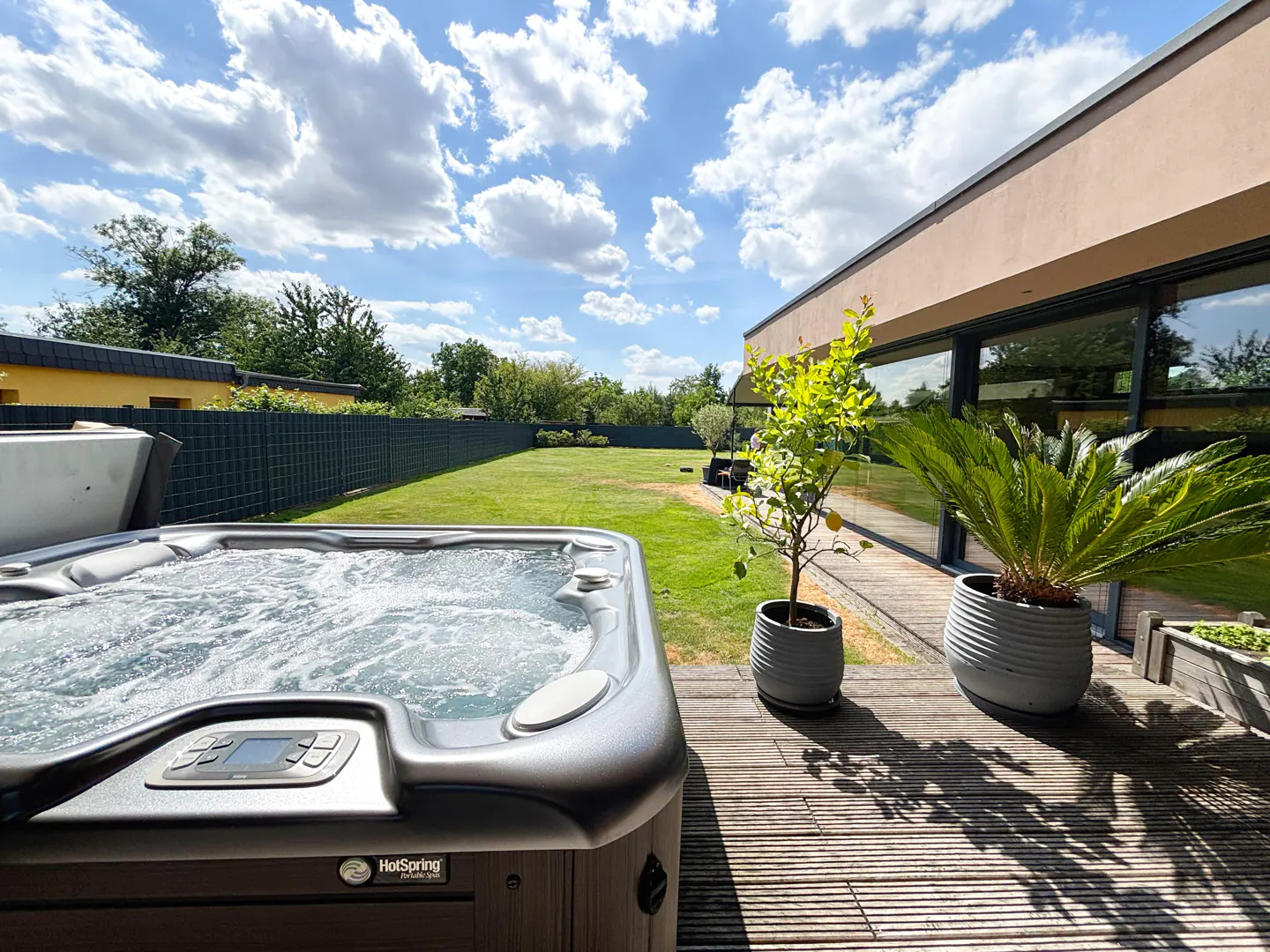 A bubbling hot tub sits on a wooden deck, overlooking a green lawn and modern house under a sunny, cloudy sky.