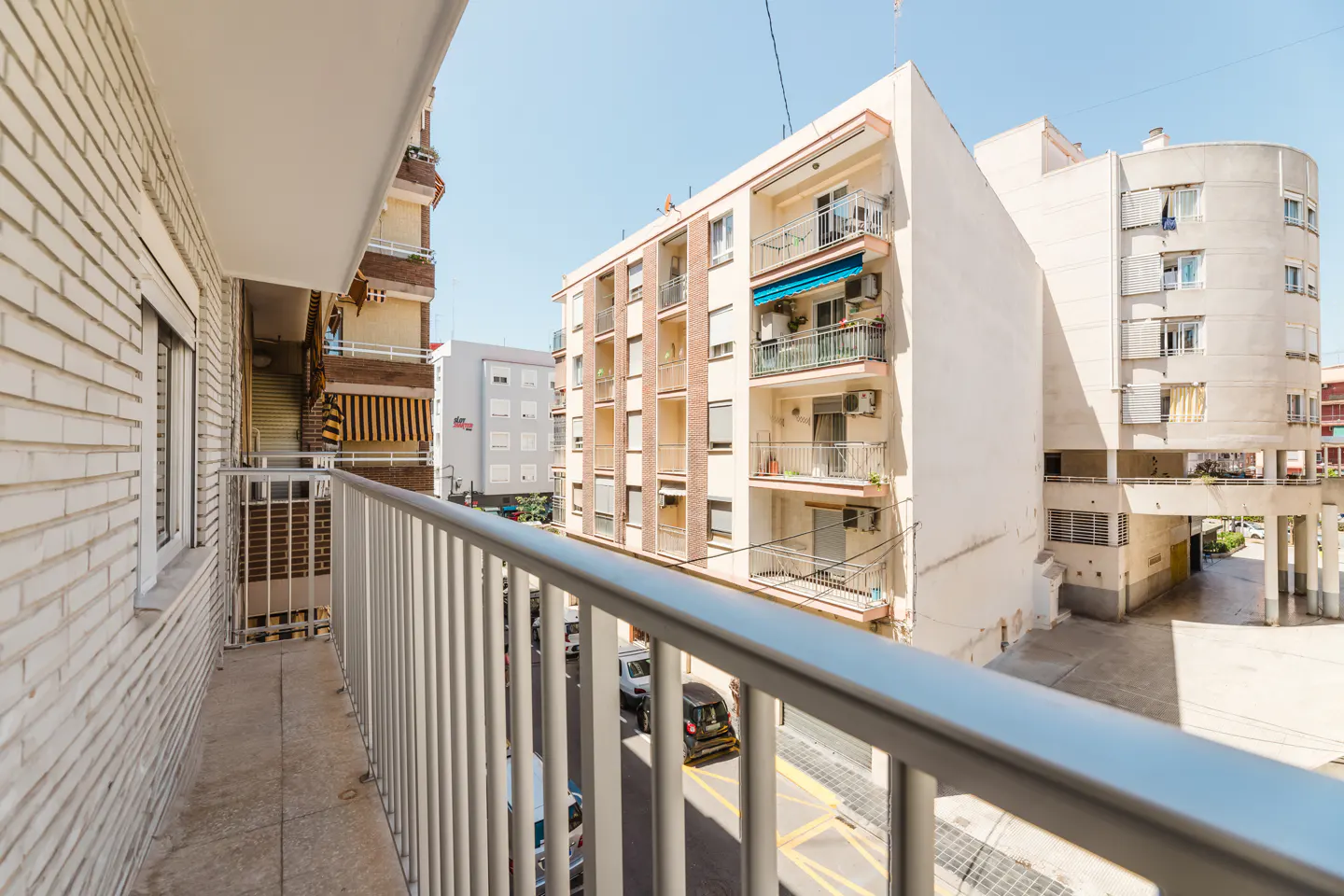 View from a balcony with a metal railing, overlooking a street with parked cars and apartment buildings.