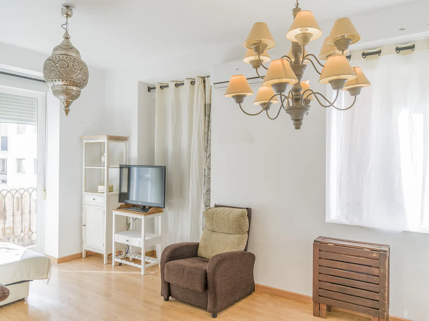 Bright living room with hardwood floors, brown armchair, TV on a white stand, and ornate chandelier and hanging lamp.