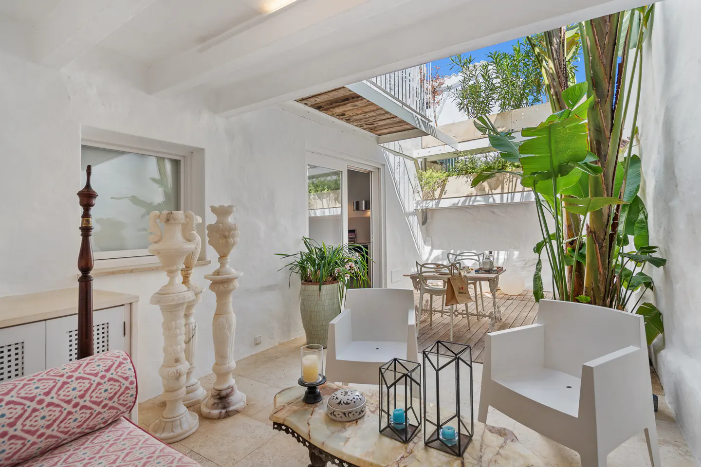 Bright patio with white walls, marble table, and white chairs. Tall green plants add a natural touch. Open roof reveals blue sky.