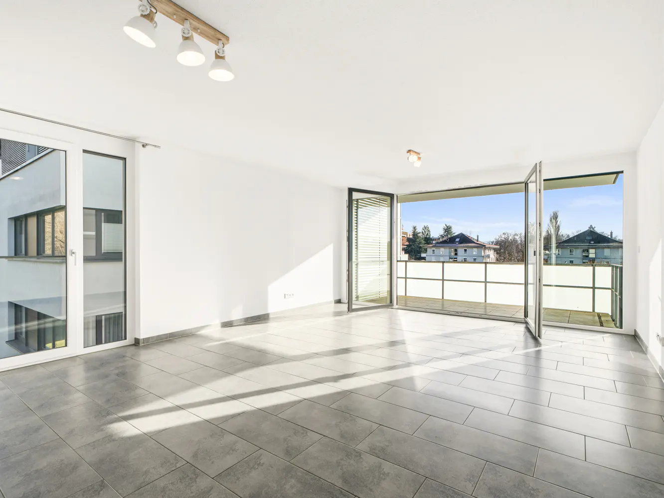 Bright, empty room with gray tile floor, white walls, and large glass doors leading to a balcony with a view of houses and trees.
