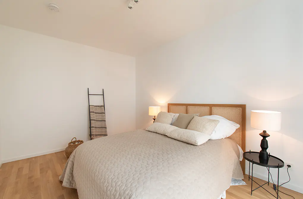 Minimalist bedroom with a beige quilted bed, rattan headboard, two bedside lamps, and a decorative ladder against the wall.