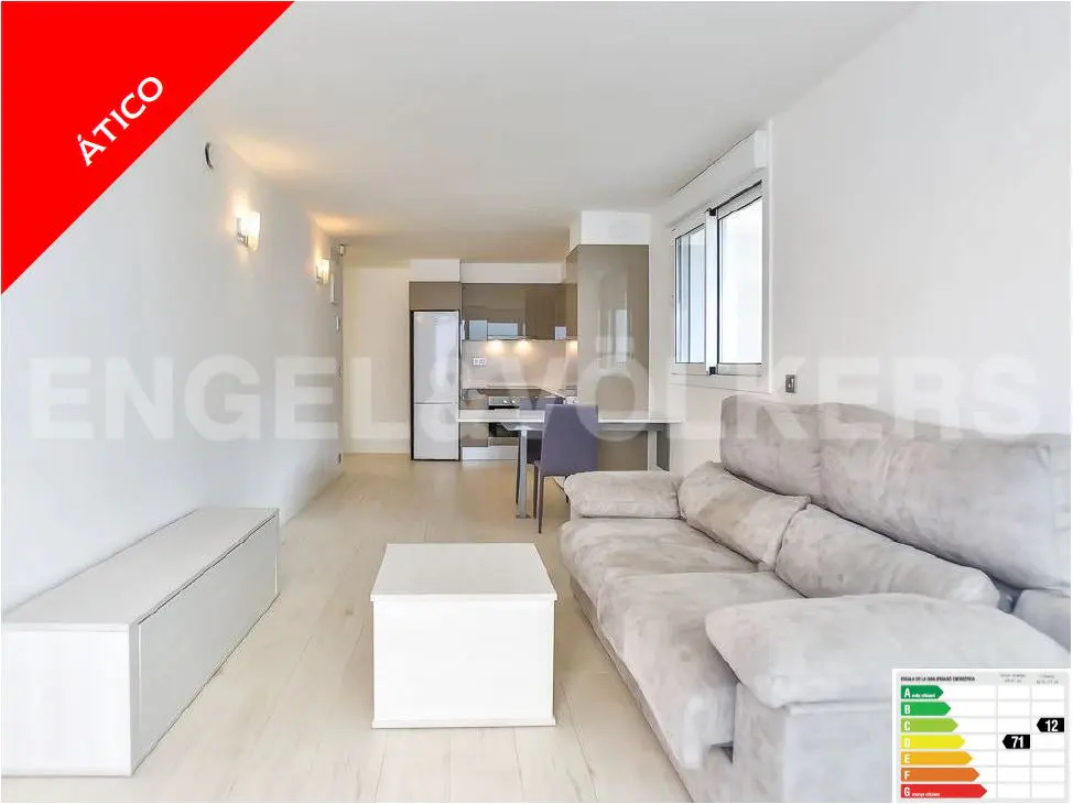 Bright, modern apartment interior with a gray sofa, white furniture, and an open kitchen. "Ático" in red on the top left.