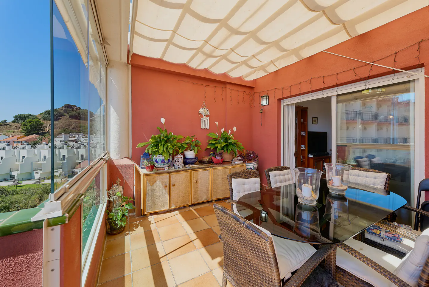 Outdoor patio with glass walls, terracotta walls, and a retractable awning. Wicker furniture surrounds a glass table. Plants decorate a wooden cabinet.