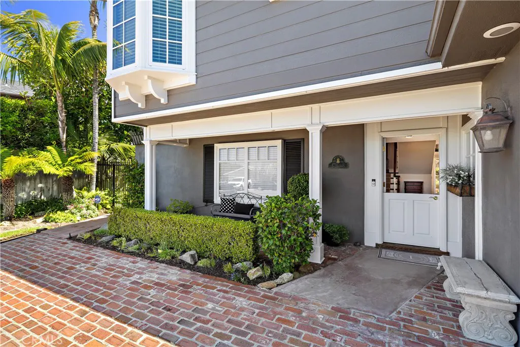 Gray house exterior with a brick patio, white Dutch door, bay window, and a small bench in a lush, sunny garden setting.