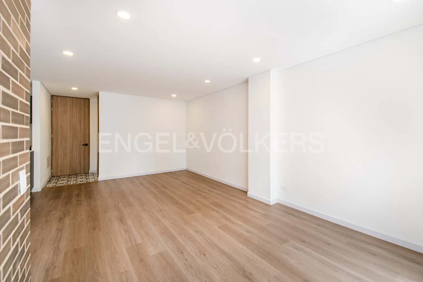 Bright, empty room with wood floors, white walls, and recessed lighting. A brick wall and wood door are on the left. Engel & Völkers logo.
