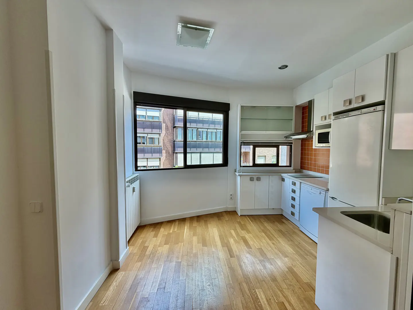 Bright kitchen with wood floors, white cabinets, and a large window. A radiator sits below the window, which overlooks a building.