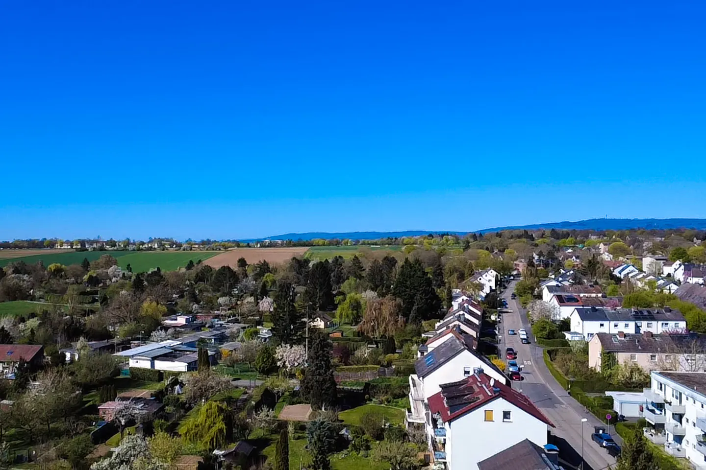 Aerial view of a suburban street with houses, trees, and cars under a clear blue sky. Fields and hills are visible in the distance.