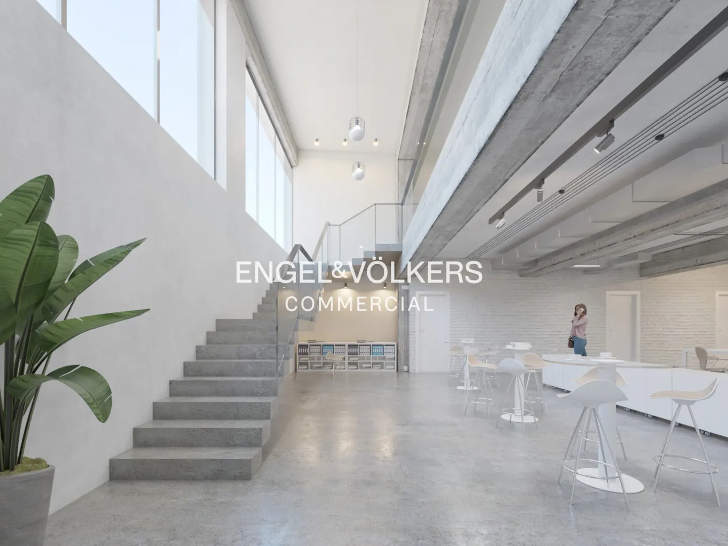 Bright, modern office space with concrete floors, stairs, and exposed ceiling beams. A large plant sits near the stairs. White bar stools line a counter.