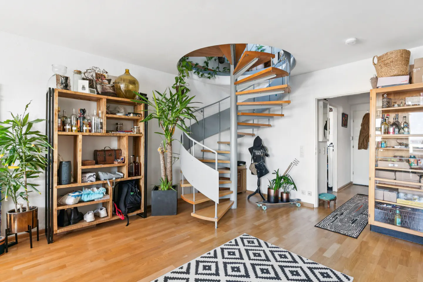 Bright living room with a spiral staircase, wooden floors, and a black and white patterned rug. Shelves display various items, and plants add greenery.
