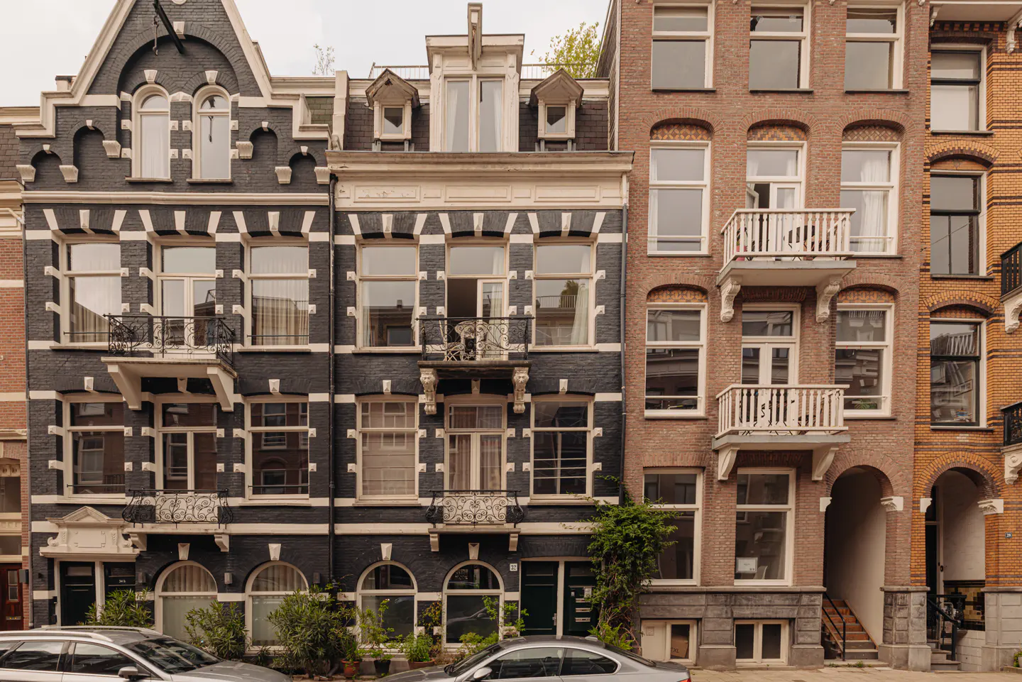 Row of narrow, multi-story brick buildings with windows and small balconies. Cars are parked in front.