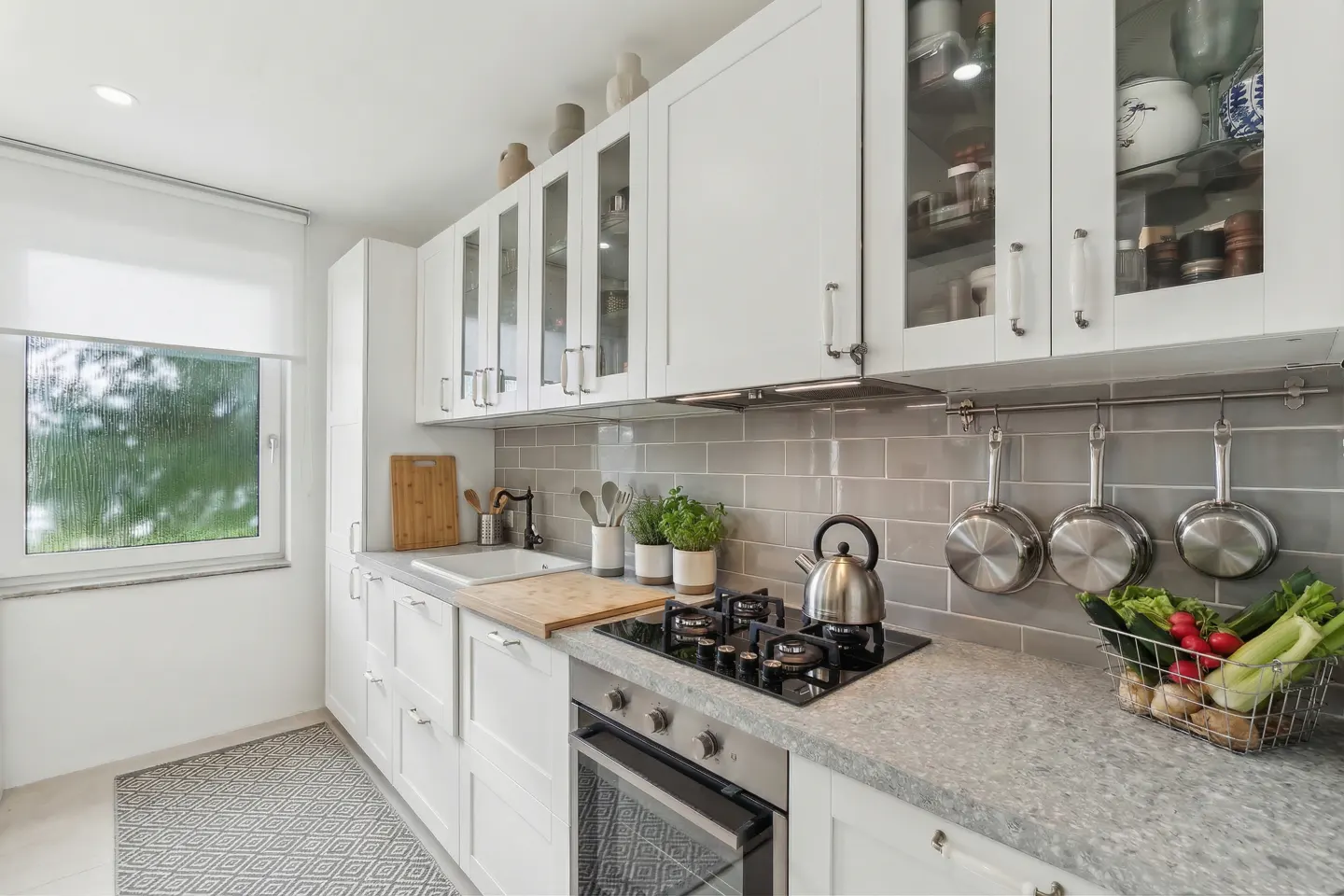 Bright kitchen with white cabinets, gray backsplash, and stainless steel appliances. A window overlooks greenery. A basket of vegetables sits on the counter.