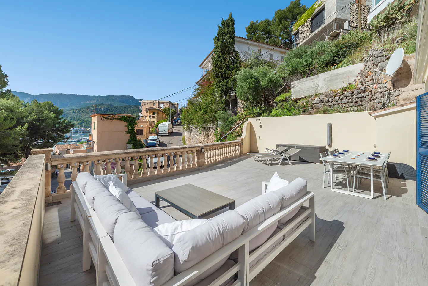 Outdoor patio with white sofa, table, and chairs. Balcony overlooks a town with mountains in the background on a sunny day.