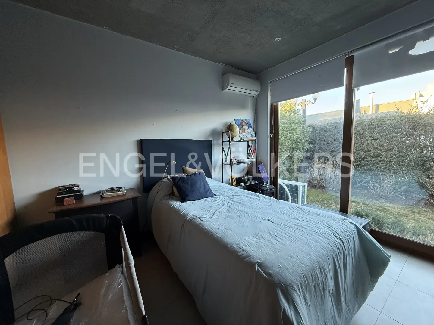 A bedroom with a bed, nightstand, and large window overlooking a green yard. The walls are light gray.