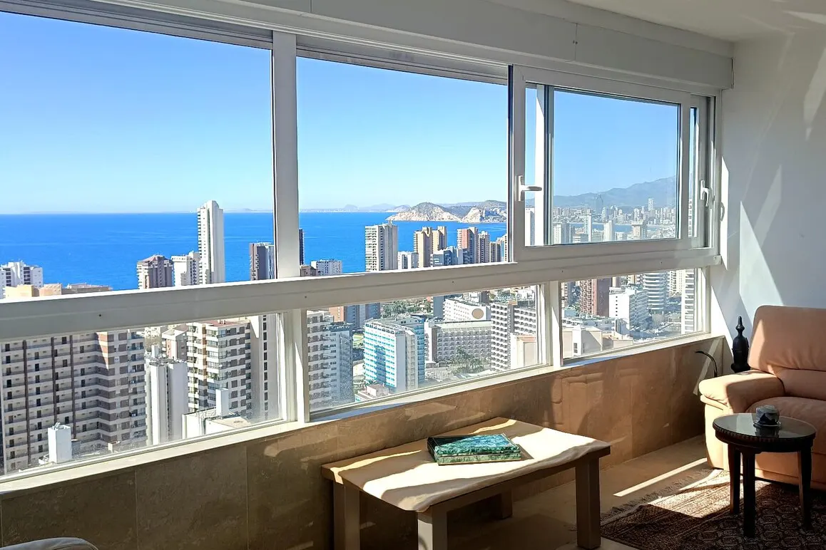 Bright apartment view through large windows overlooking the city, ocean, and mountains. A small table and chair sit in the foreground.