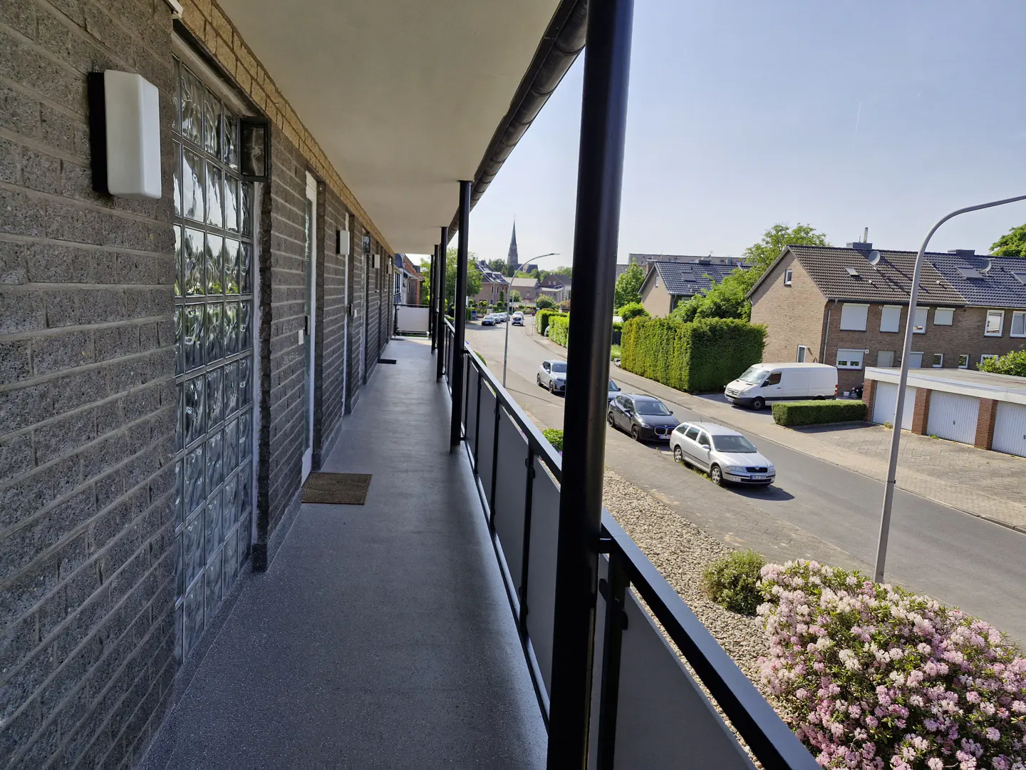 A long, covered balcony with a grey brick wall and glass block windows overlooking a quiet suburban street with cars.