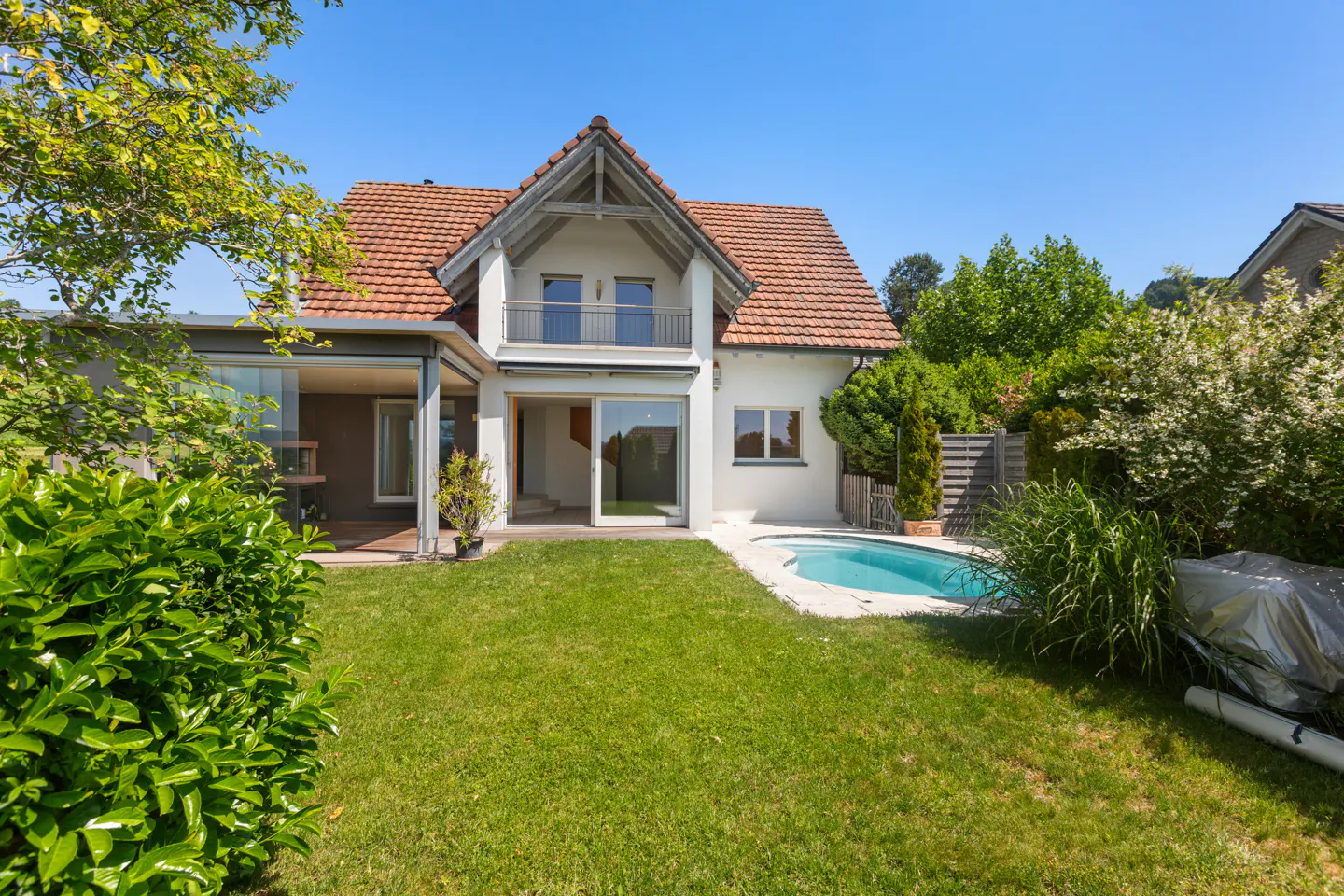 Backyard of a white two-story house with a red tile roof, a small swimming pool, and a lush green lawn under a blue sky.