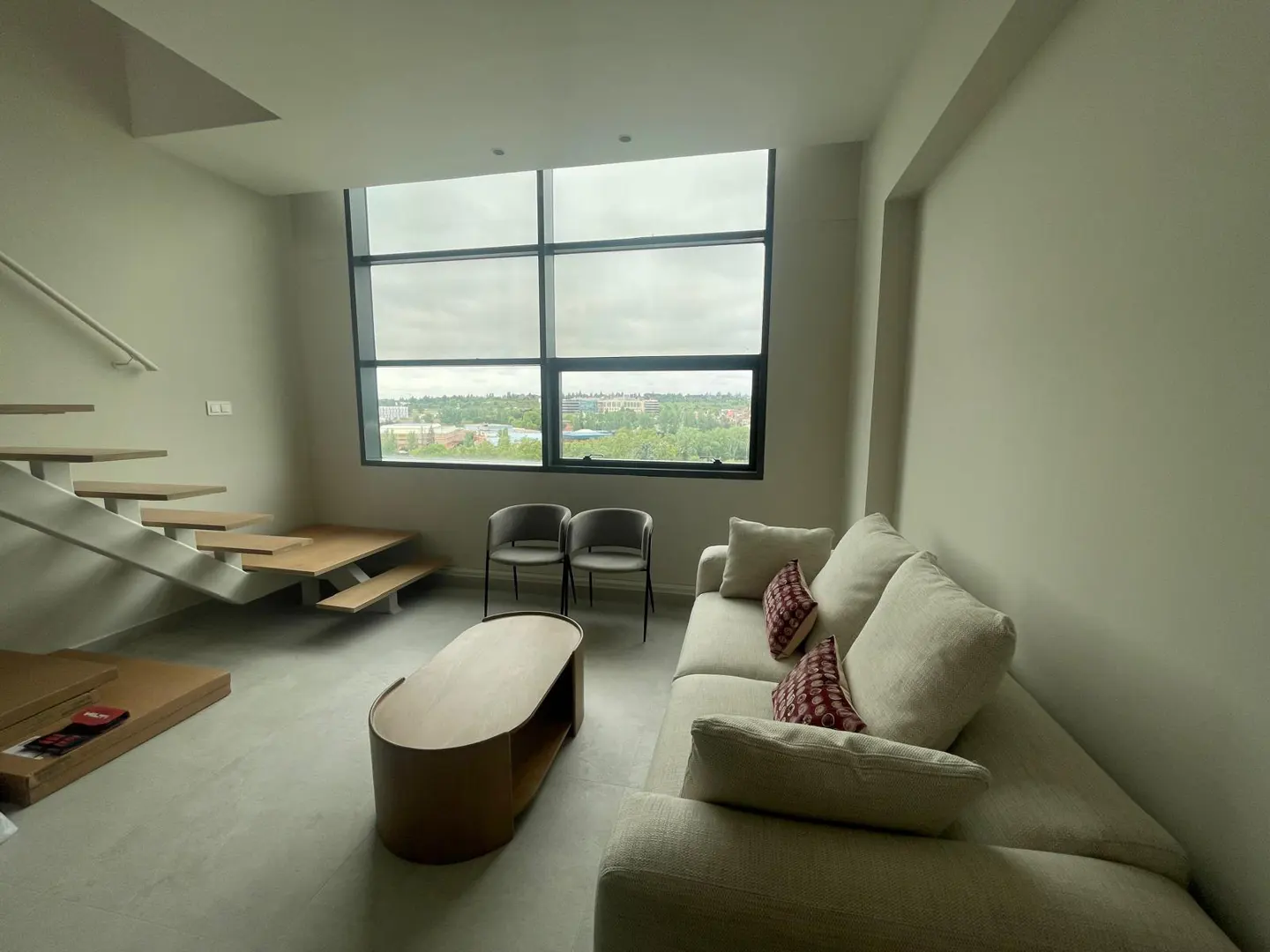 Living room with a white sofa, wood coffee table, and two chairs near a large window with a city view. Stairs are on the left.