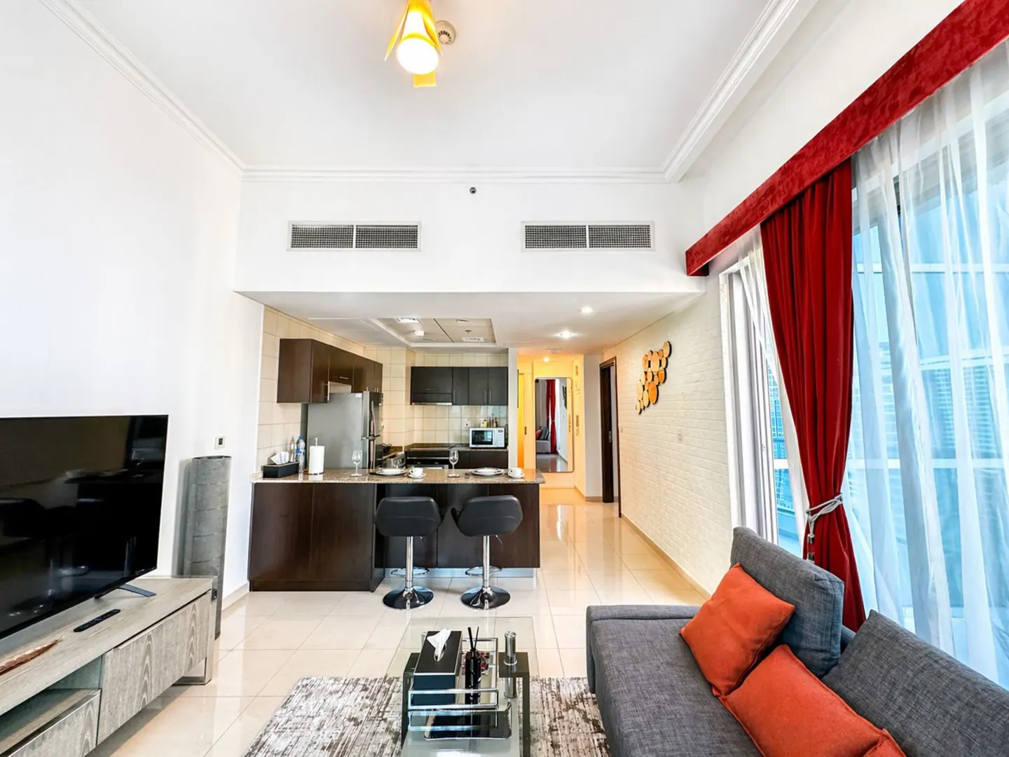 Bright, modern apartment interior with a gray sofa, orange pillows, and a kitchen with dark wood cabinets and bar stools. Red curtains frame a large window.