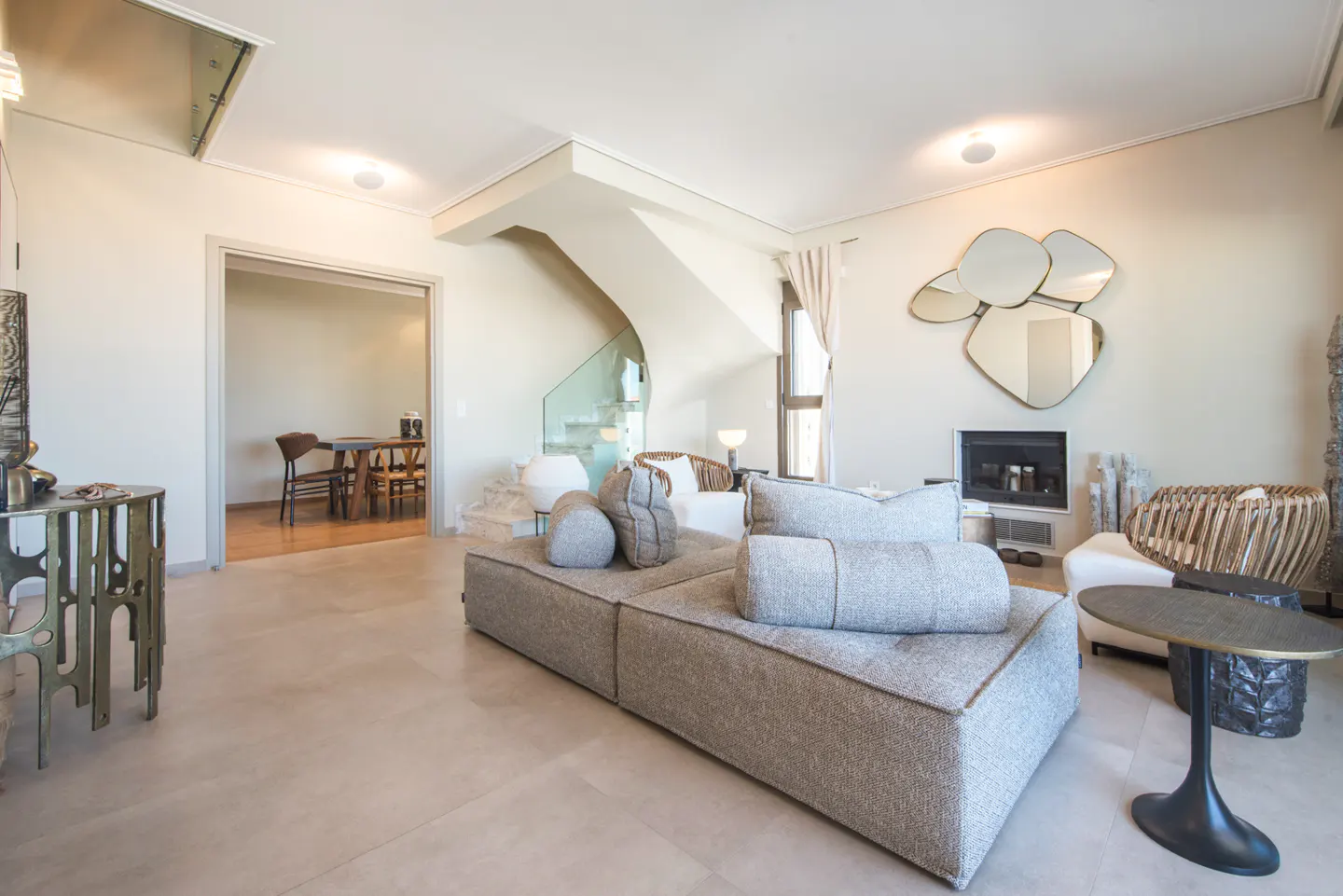 Bright living room with a gray modular sofa, fireplace, and unique mirrors. A dining area is visible through an open doorway.