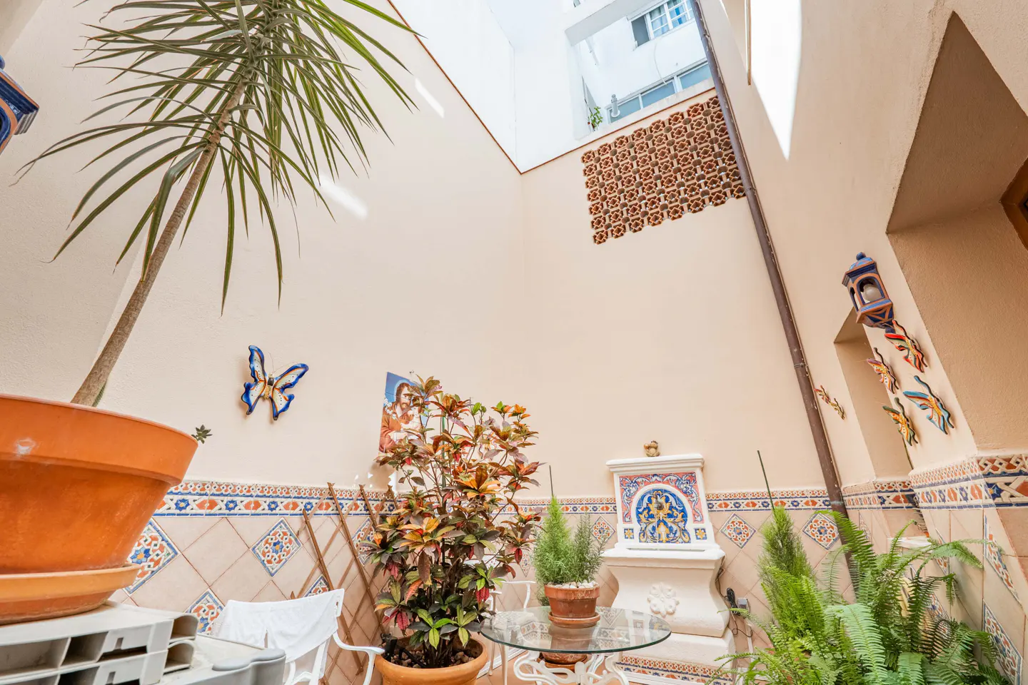 A sunlit interior courtyard with beige walls, decorative tiles, potted plants, a small fountain, and butterfly wall art.