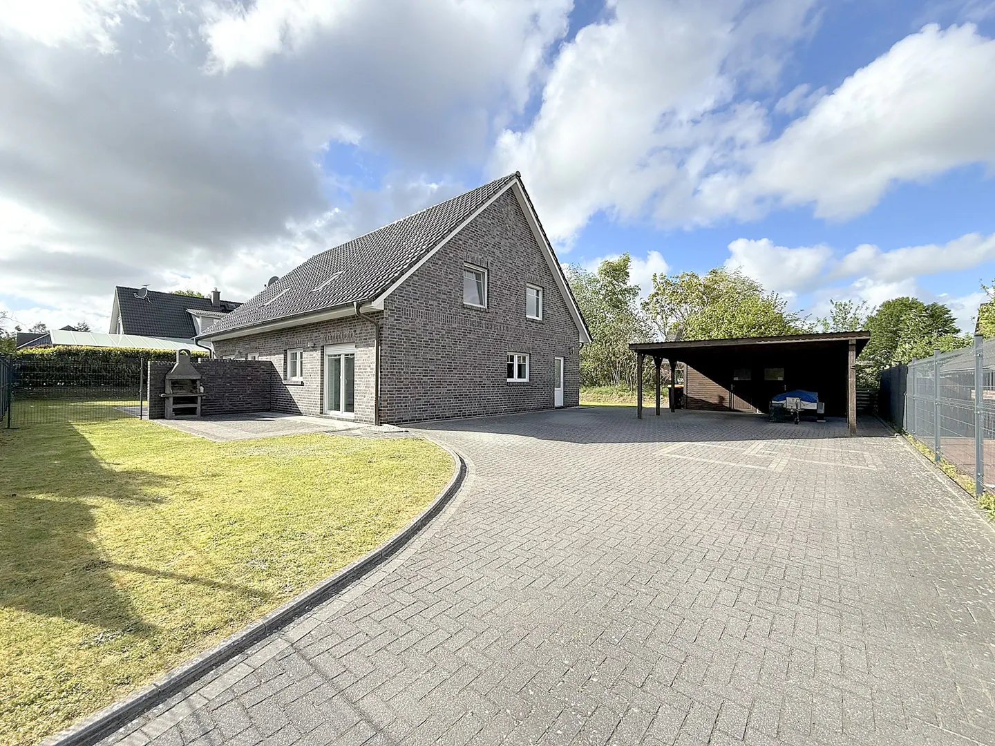 Gray brick house with a paved driveway, a carport, and a green lawn under a cloudy blue sky.