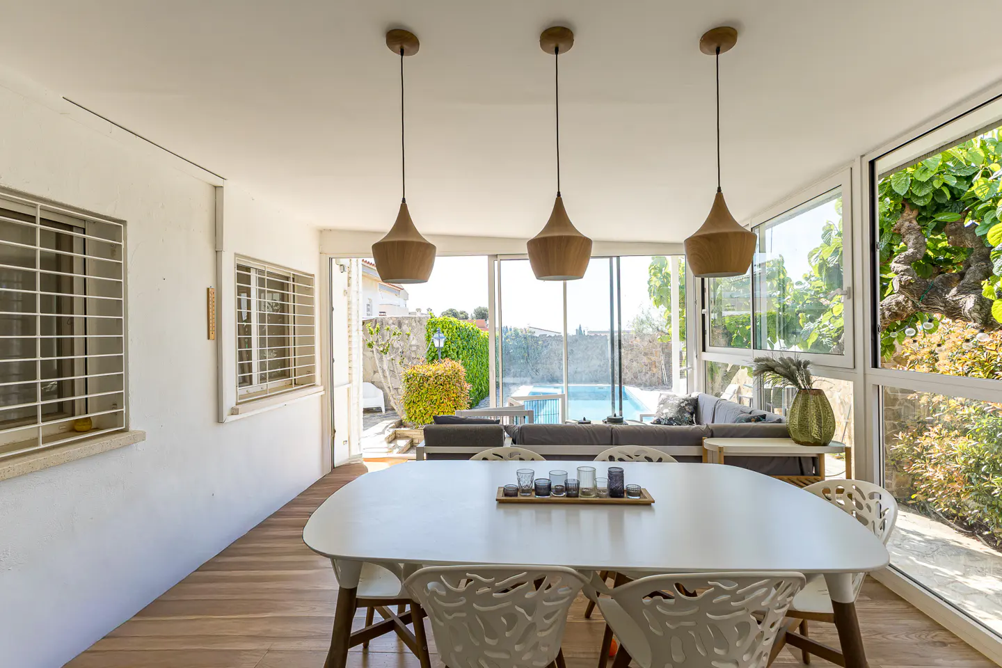 Bright sunroom with a white table, chairs, and pendant lights. A pool and greenery are visible through the windows.