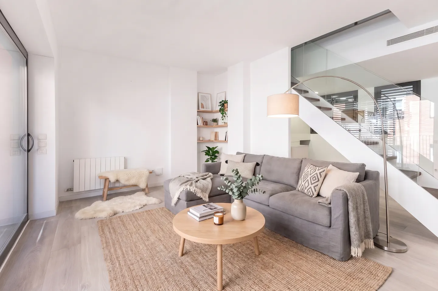 Bright living room with a gray sectional sofa, round wood table, and jute rug. White walls, wood floors, and stairs with glass railing.