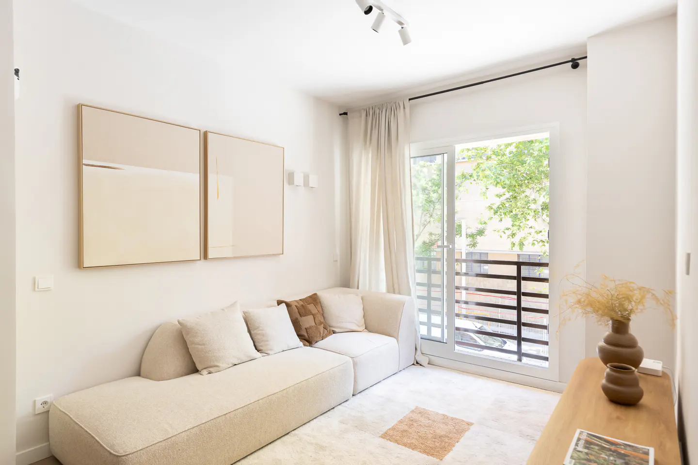 Bright living room with a beige modular sofa, two abstract wall prints, and a balcony door overlooking green trees.