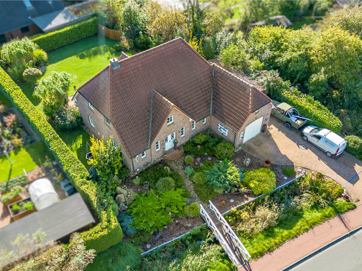 Aerial view of a brick house with a brown roof, surrounded by green lawns, trees, and a driveway with two parked vehicles.