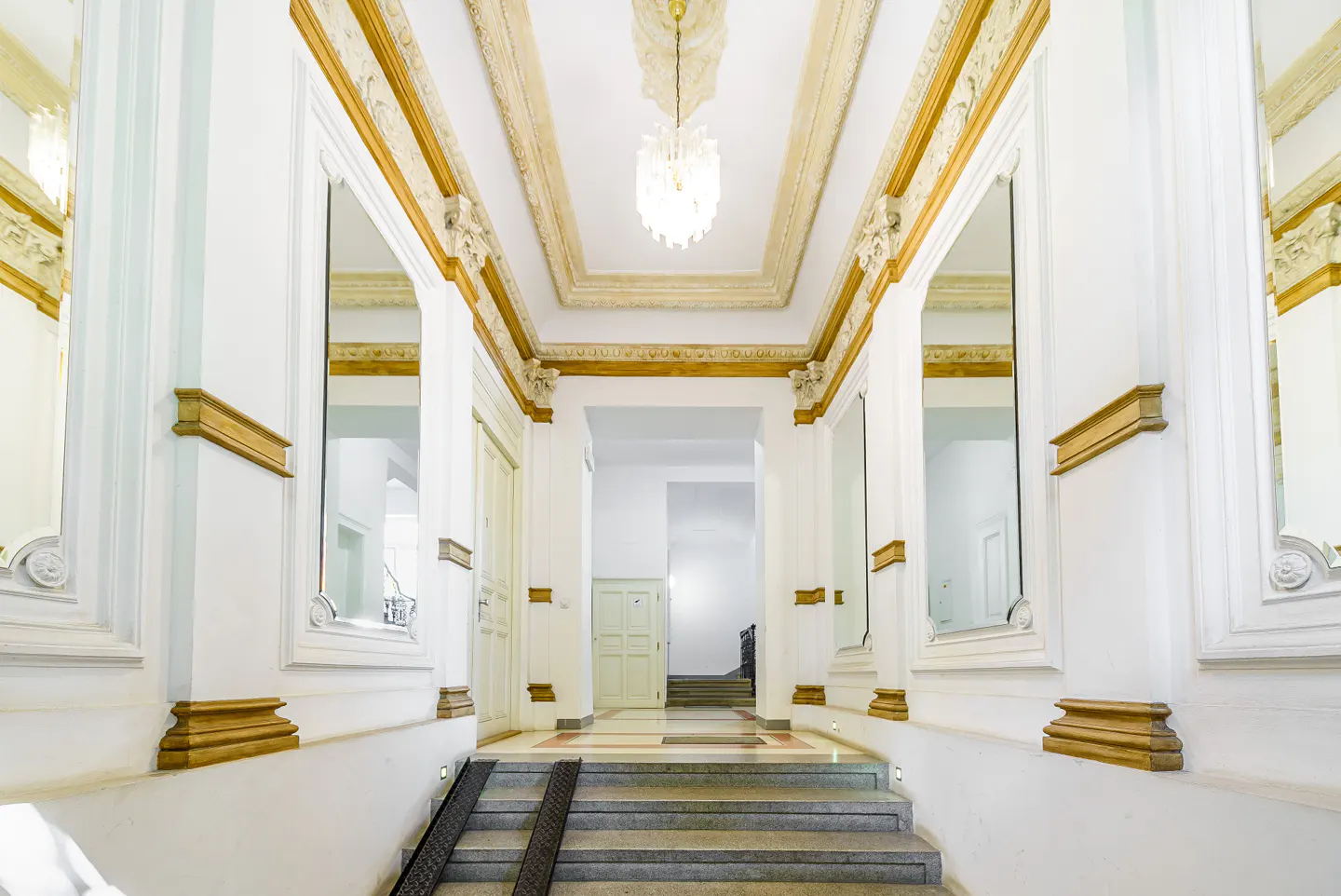 Ornate white hallway with gold trim, mirrors, and a crystal chandelier. Steps lead to a doorway. Ramp access is available.