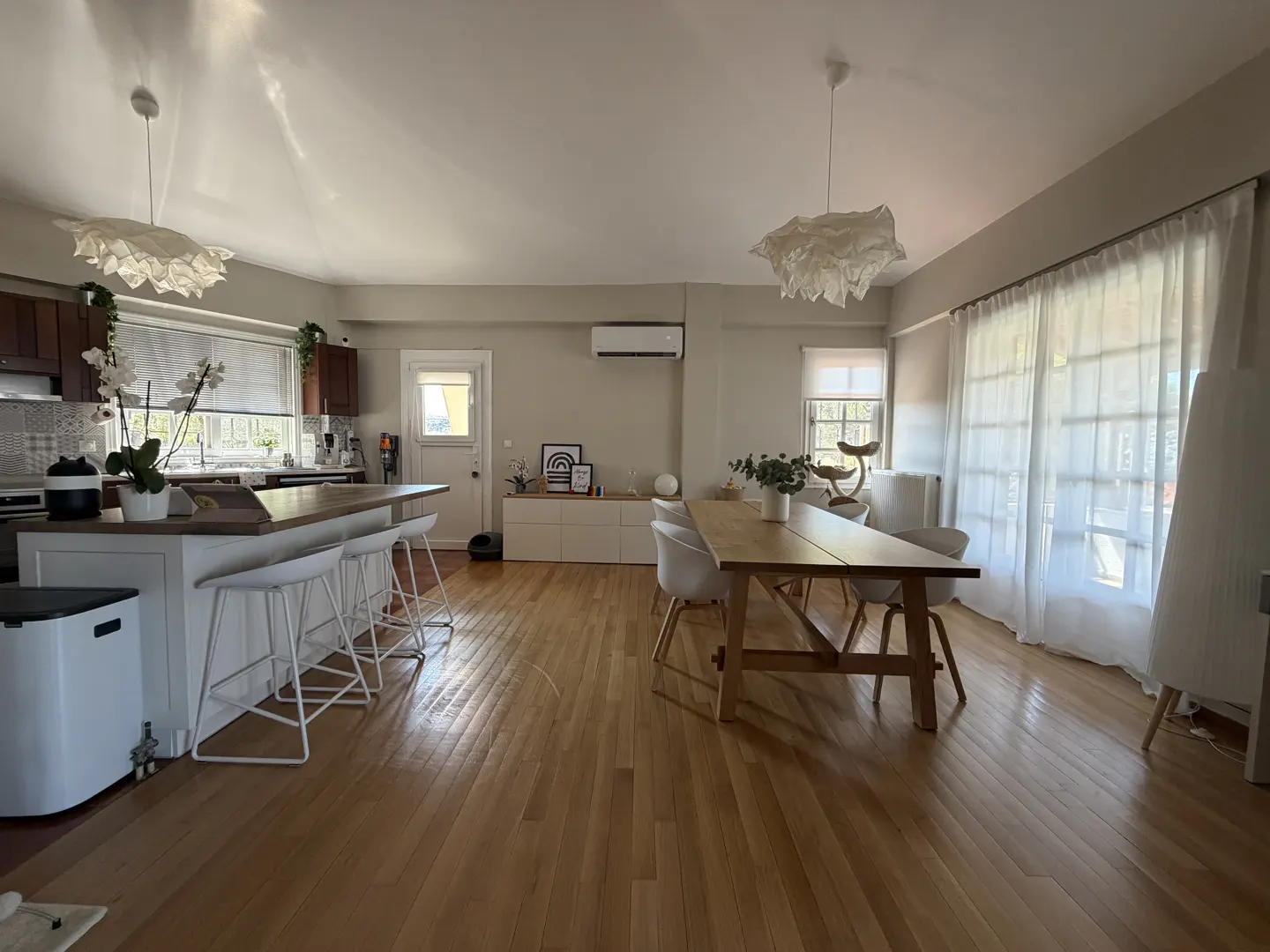 Open-concept kitchen and dining area with hardwood floors, white bar stools, a wooden dining table, and sheer curtains.