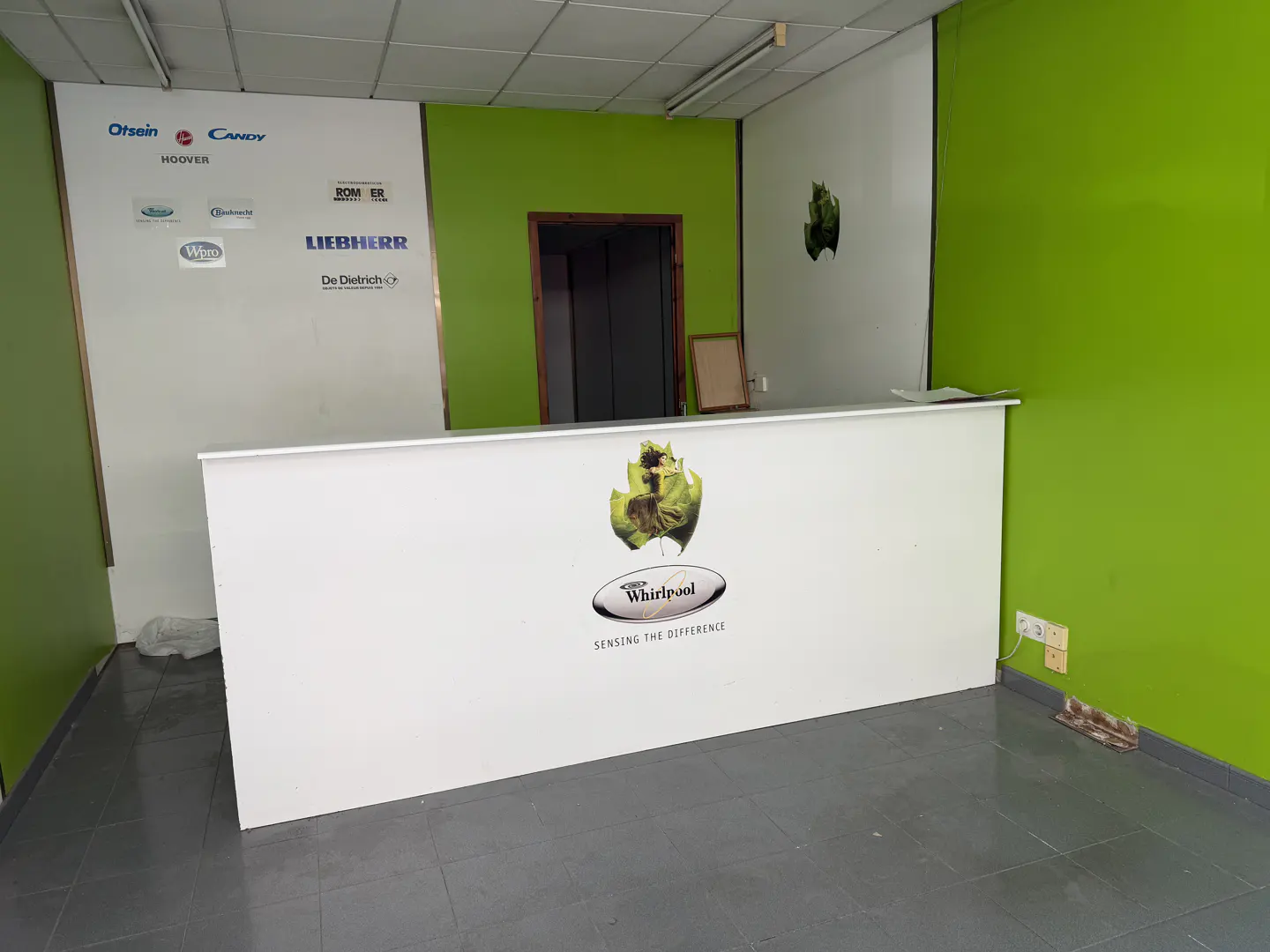 Interior view of a commercial space with a white reception desk featuring the Whirlpool logo, green accent walls, and appliance brand logos on the wall.