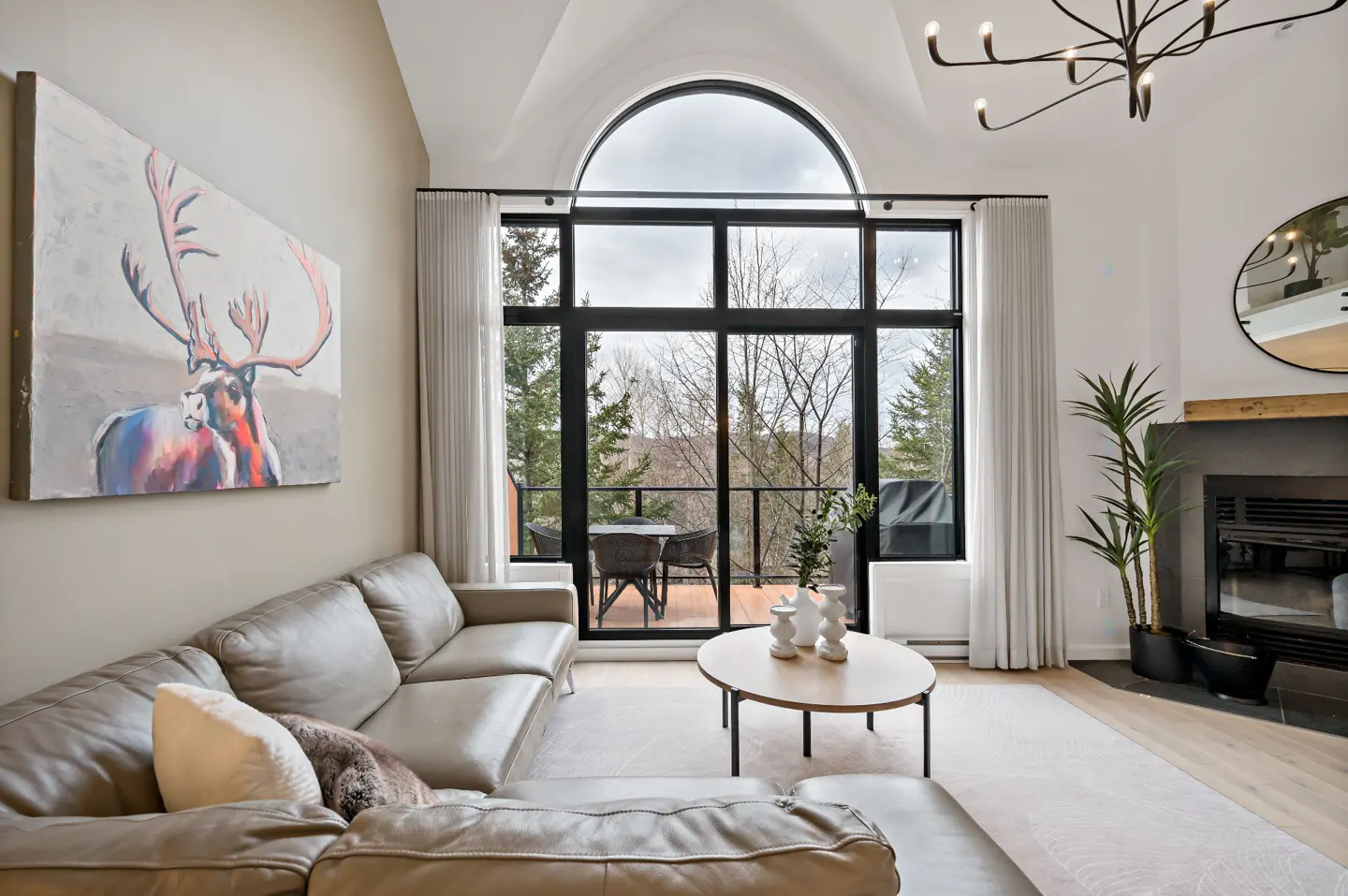 Living room with a gray leather sofa, a painting of a deer, a fireplace, and a large window with a view of trees.