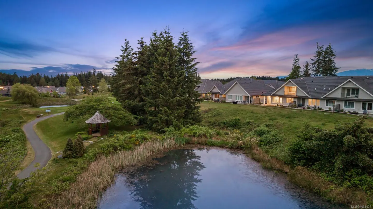 Aerial view of townhouses with a pond, gazebo, and walking path at dusk. The sky is blue and pink.
