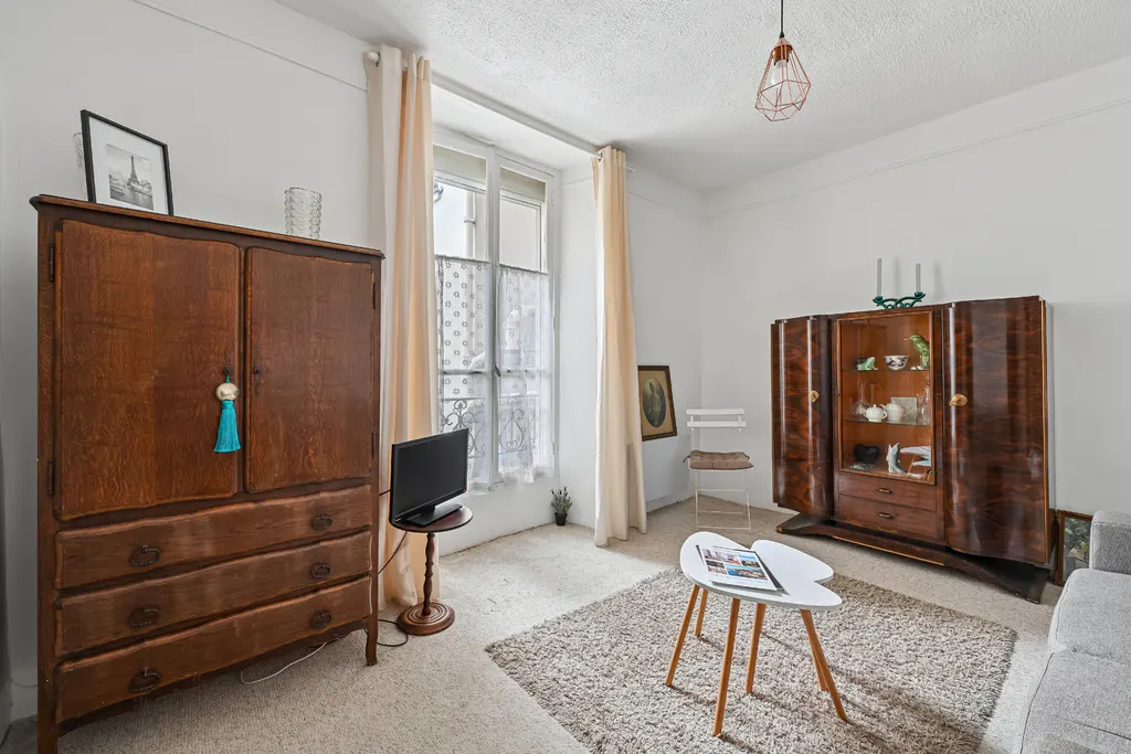Bright living room with white walls, a window with sheer curtains, and vintage furniture including a wooden armoire and display cabinet.