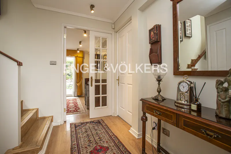 Hallway with light wood floors, white walls, and a patterned rug. A console table with a lamp and clock sits near a mirror reflecting stairs.