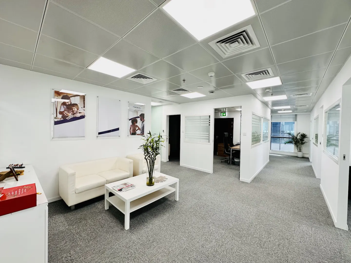 Bright office lobby with white walls, gray carpet, and a white sofa and coffee table. Posters hang on the wall.