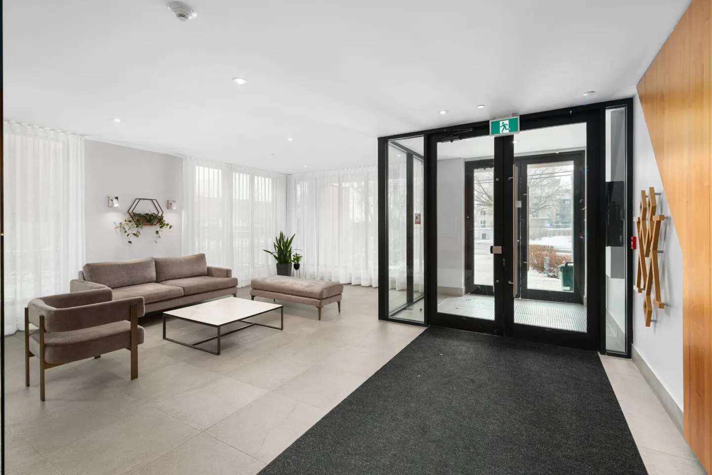Modern apartment lobby with a grey sofa, armchair, coffee table, and large glass entrance doors with a black frame.