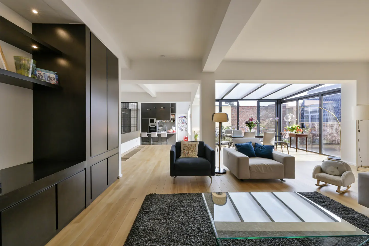 Bright, open living room with hardwood floors, black built-ins, and a glass-enclosed sunroom. Furniture includes a gray sofa with blue pillows.
