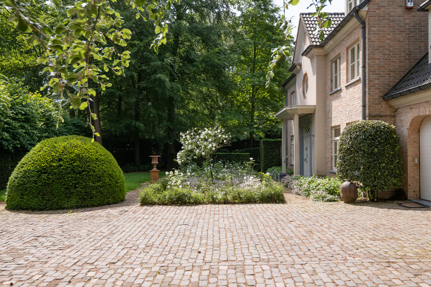 Brick driveway leads to a brick house with a light blue door. Green trees and bushes surround the house.