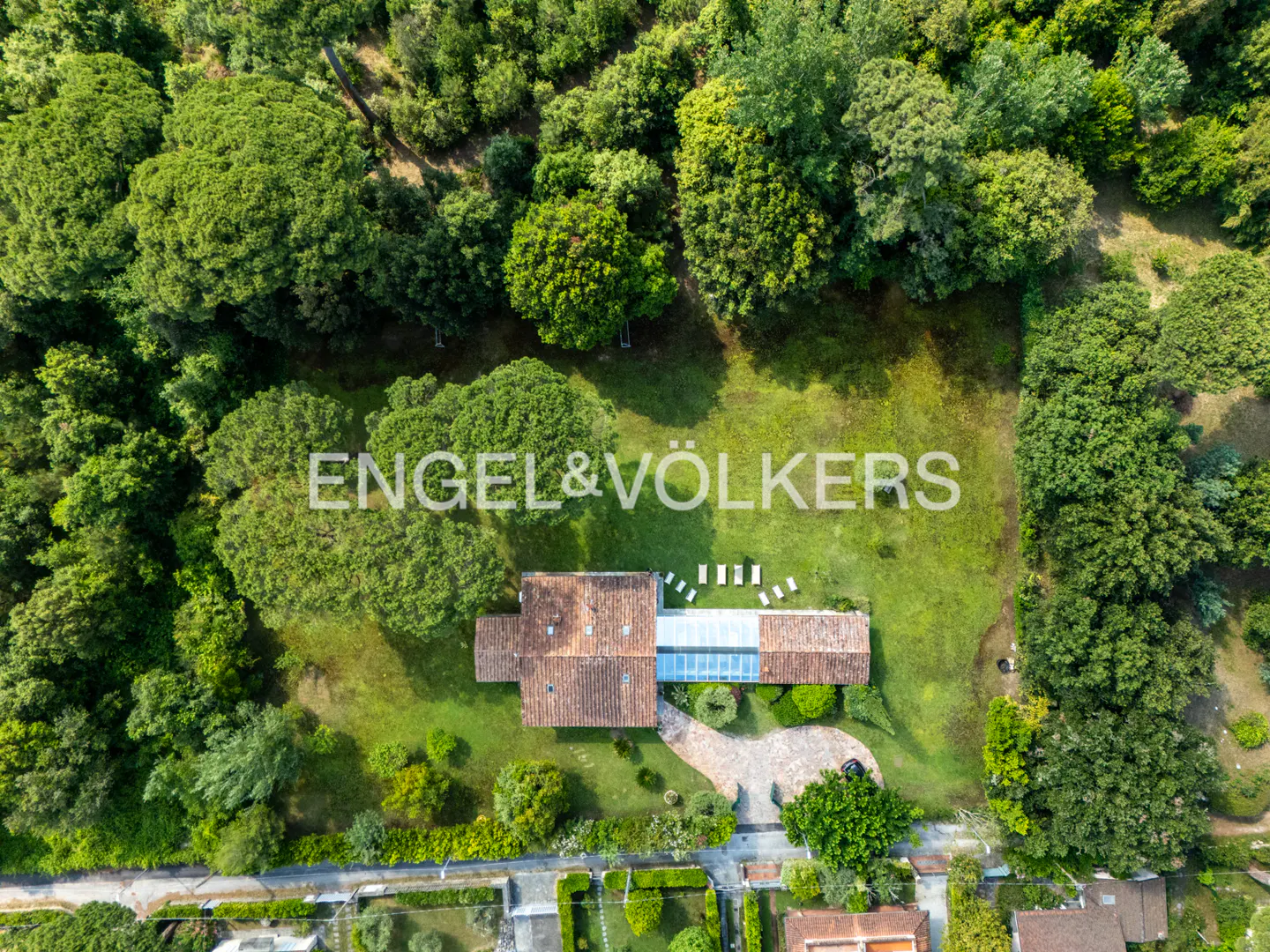 Aerial view of a house with a tiled roof and glass sunroom, surrounded by a large green lawn and dense forest trees.