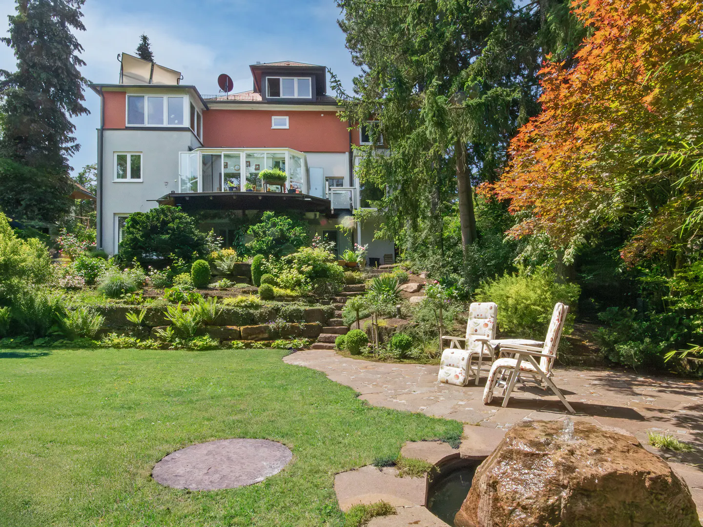 A gray and red house with a lush green garden, stone steps, and two chairs on a patio.