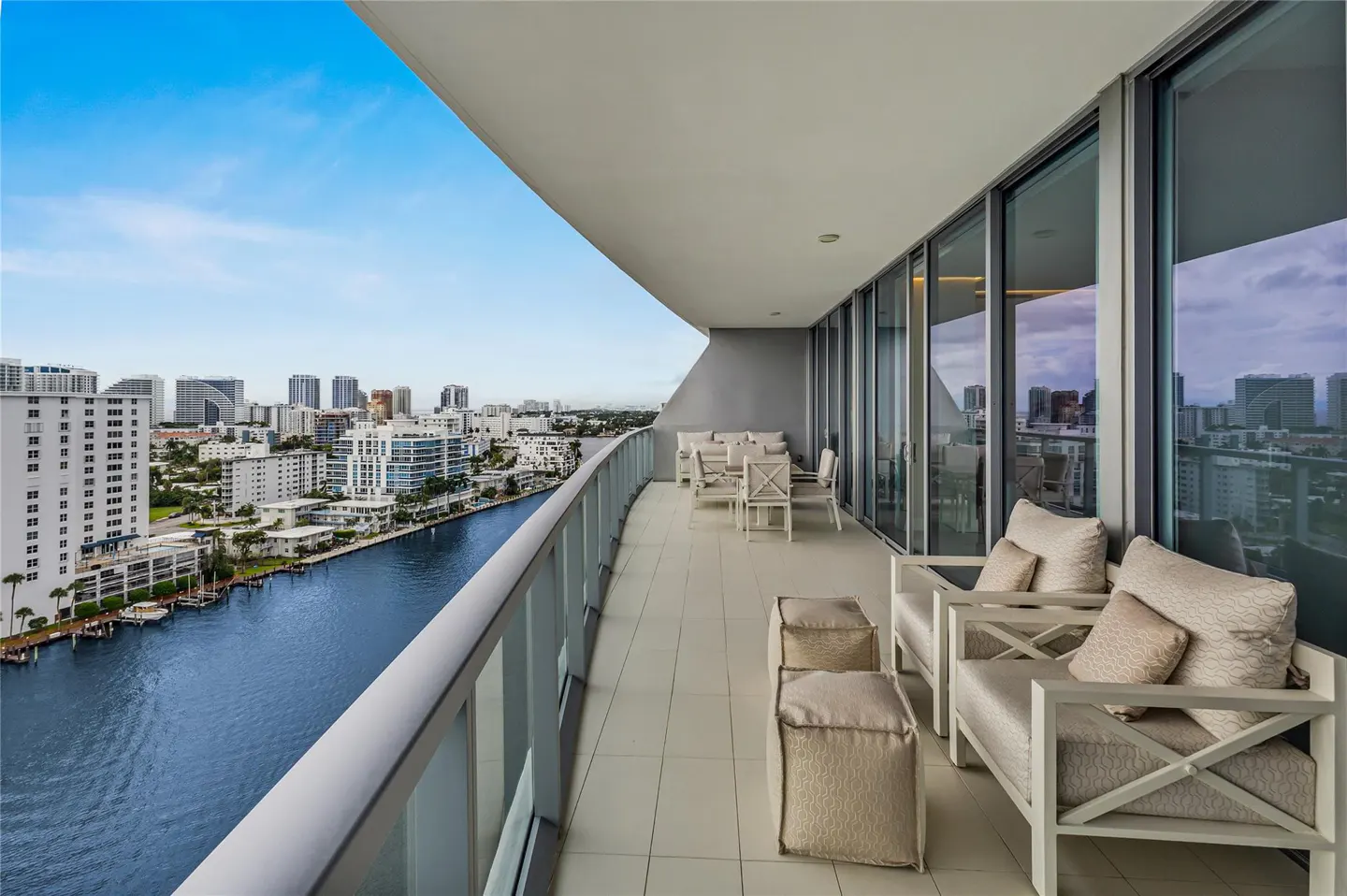 A condo balcony with white furniture overlooks a blue river and city skyline under a blue sky.